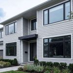 Modern two-story house with light-gray siding, large windows, and a small front canopy over the entrance; landscaped front yard with shrubs and trees