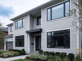 Modern two-story house with light-gray siding, large windows, and a small front canopy over the entrance; landscaped front yard with shrubs and trees
