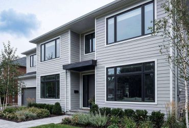 Modern two-story house with light-gray siding, large windows, and a small front canopy over the entrance; landscaped front yard with shrubs and trees