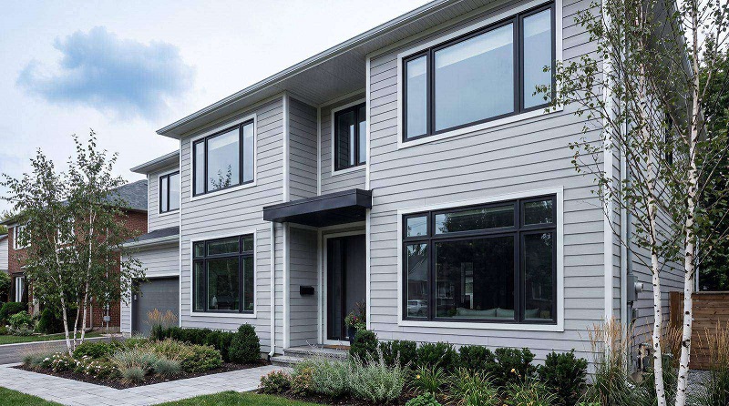 Modern two-story house with light-gray siding, large windows, and a small front canopy over the entrance; landscaped front yard with shrubs and trees