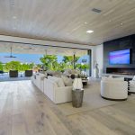 Spacious modern living room with white sofas, a fireplace, and a large wall-mounted TV, open to a bright outdoor patio.