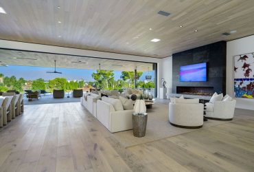 Spacious modern living room with white sofas, a fireplace, and a large wall-mounted TV, open to a bright outdoor patio.