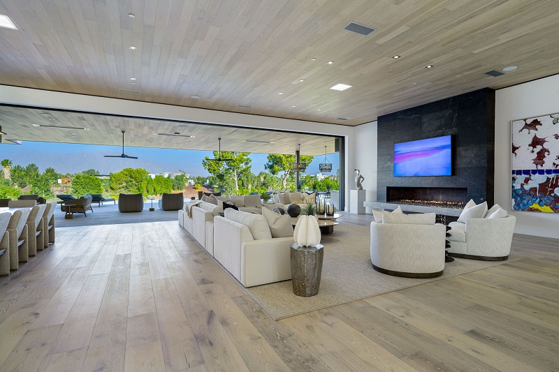 Spacious modern living room with white sofas, a fireplace, and a large wall-mounted TV, open to a bright outdoor patio.