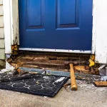 Front door with blue panel and damaged threshold; exposed wood and tools (hammer) on a mat outside the doorway.