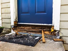 Front door with blue panel and damaged threshold; exposed wood and tools (hammer) on a mat outside the doorway.