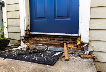Front door with blue panel and damaged threshold; exposed wood and tools (hammer) on a mat outside the doorway.