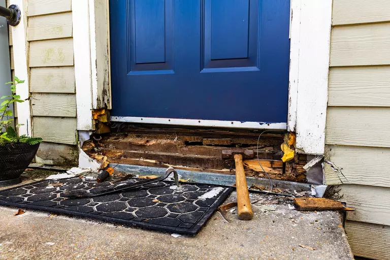 Front door with blue panel and damaged threshold; exposed wood and tools (hammer) on a mat outside the doorway.