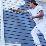 Man in a white shirt and cap spraying blue paint onto the exterior siding of a house.
