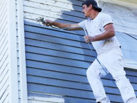 Man in a white shirt and cap spraying blue paint onto the exterior siding of a house.