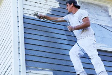 Man in a white shirt and cap spraying blue paint onto the exterior siding of a house.