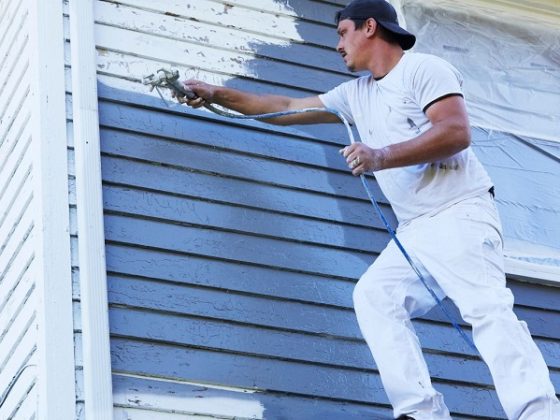 Man in a white shirt and cap spraying blue paint onto the exterior siding of a house.
