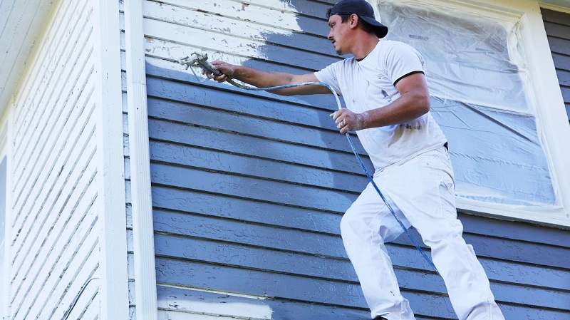 Man in a white shirt and cap spraying blue paint onto the exterior siding of a house.