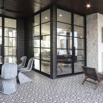 Covered outdoor patio with glass walls and black metal frames, featuring a round wicker dining table and six chairs on a patterned tile floor, plus a fireplace corner with lounge chairs nearby.