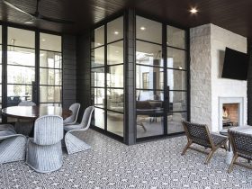 Covered outdoor patio with glass walls and black metal frames, featuring a round wicker dining table and six chairs on a patterned tile floor, plus a fireplace corner with lounge chairs nearby.