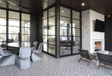 Covered outdoor patio with glass walls and black metal frames, featuring a round wicker dining table and six chairs on a patterned tile floor, plus a fireplace corner with lounge chairs nearby.