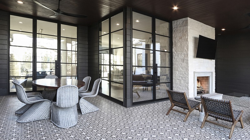 Covered outdoor patio with glass walls and black metal frames, featuring a round wicker dining table and six chairs on a patterned tile floor, plus a fireplace corner with lounge chairs nearby.
