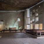 Interior of a modern church/chapel with a raised altar area left, a simple cross, and rows of wooden pews on the right under a curved ceiling and geometric wall openings