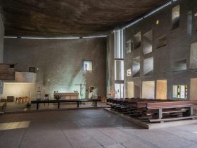 Interior of a modern church/chapel with a raised altar area left, a simple cross, and rows of wooden pews on the right under a curved ceiling and geometric wall openings