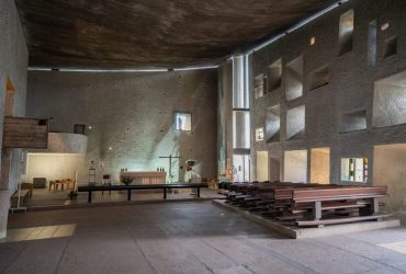 Interior of a modern church/chapel with a raised altar area left, a simple cross, and rows of wooden pews on the right under a curved ceiling and geometric wall openings