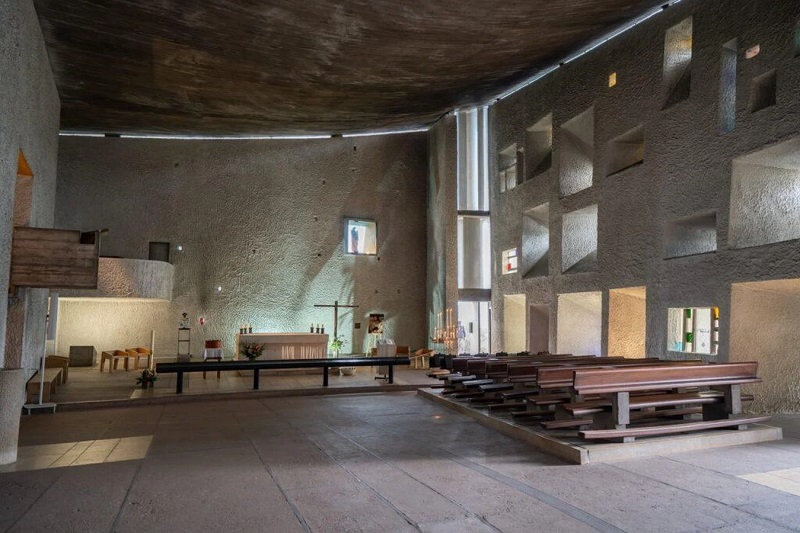Interior of a modern church/chapel with a raised altar area left, a simple cross, and rows of wooden pews on the right under a curved ceiling and geometric wall openings